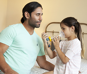 Man helping girl use metered-dose inhaler with spacer and mask.